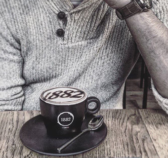 Mann sitzt am Tisch und hat einen Cappuccino mit der Jahreszahl 1882 als Latte Art vor sich