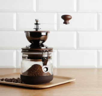 Hario Handmühle Canister auf einem Tablett mit Kaffeebohnen vor einer weißen Fliesenwand