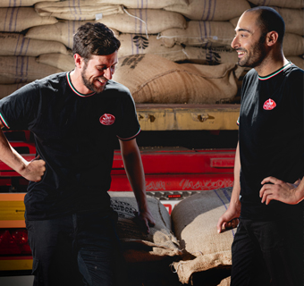 Zwei Personen mit Saquella Logo T-Shirt vor Kaffeesäcken