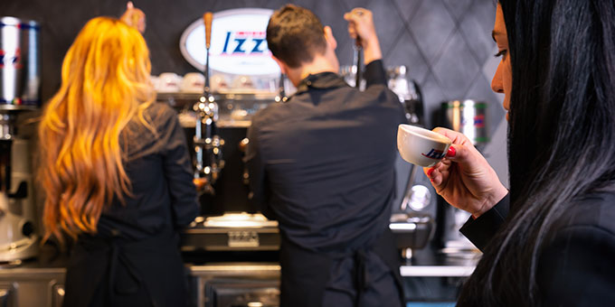 Im Hintergrund stehen zwei Personen an einer Kaffeemaschine, im Vordergrund trinkt eine Person Espresso