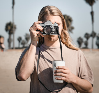 Person fotografiert mit einer Kamera dabei hält sie weißen Thermobecher in der anderen Hand